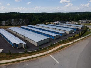 Aerial view of 60x100 mini storage steel buildings with blue roll-up doors, designed for efficient storage solutions, surrounded by greenery and paved access roads.