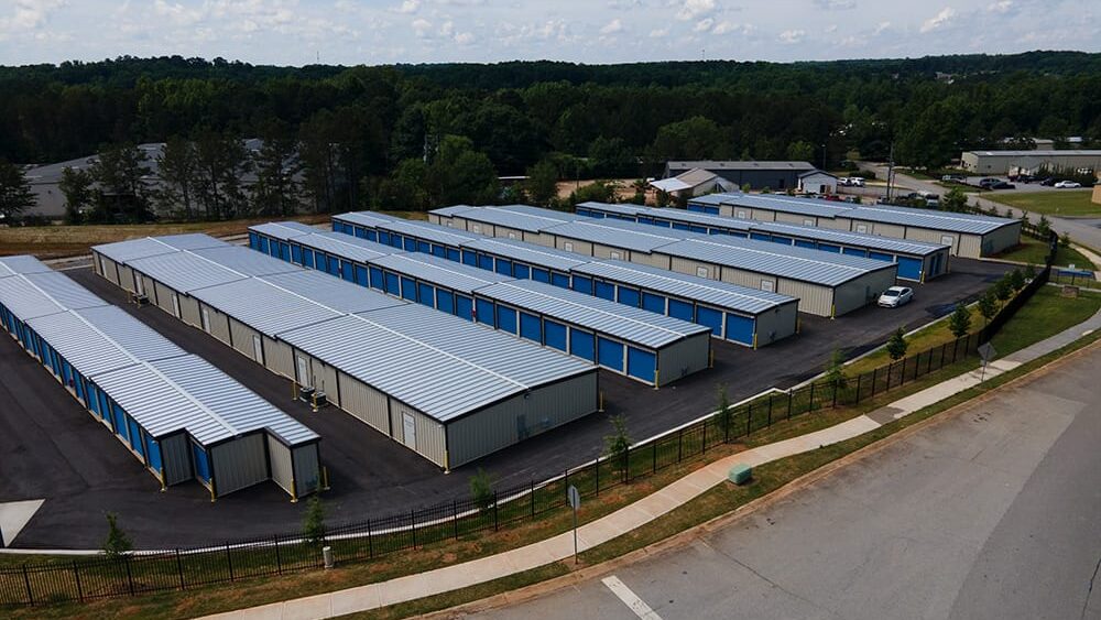 Aerial view of 60x100 mini storage steel buildings with blue roll-up doors, designed for efficient storage solutions, surrounded by greenery and paved access roads.